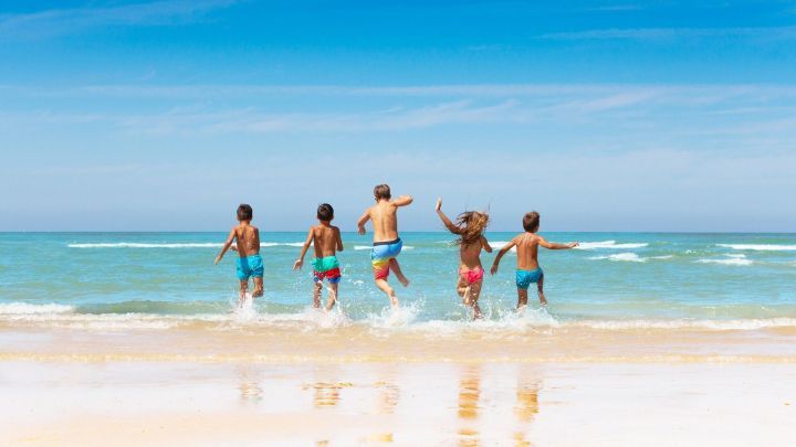 F&uuml;nf Kinder laufen fr&ouml;hlich ins Meer und spritzen Wasser, w&auml;hrend sie am sonnigen Strand spielen.