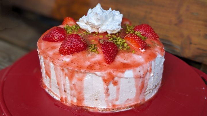 Eine kleine Geburtstagstorte mit Erdbeeren steht auf einem roten Teller, dekoriert mit Sahne und Pistazien, vor einem Holzhintergrund.