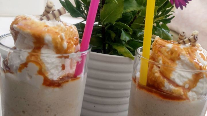 Zwei Gl&auml;ser Frappuccino mit Karamell und Sahne stehen auf einem Tisch, umgeben von einer bl&uuml;henden Pflanze.