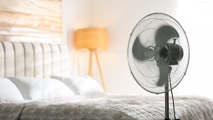 Ein Ventilator steht in einem k&uuml;hlen Schlafzimmer und bl&auml;st frische Luft &uuml;ber das Bett, um Sommerhitze zu bek&auml;mpfen.