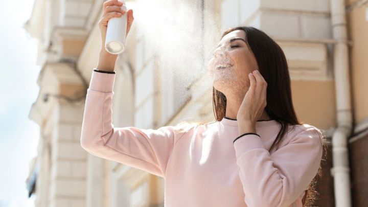 Eine Frau spr&uuml;ht ein k&uuml;hlendes Spray auf ihr Gesicht, w&auml;hrend sie in der warmen Stadt an einem sonnigen Tag steht.