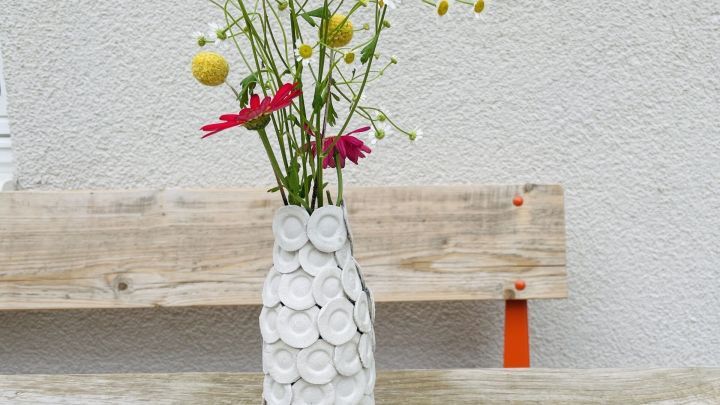 Eine Strukturvase aus Eierkarton h&auml;lt bunte Blumen, w&auml;hrend sie auf einer Holzbank vor einer grauen Wand steht.