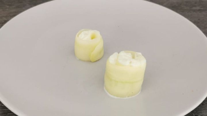 Auf einem wei&szlig;en Teller liegen zwei Sushi-Rollen aus Gurke, gef&uuml;llt mit einer cremigen F&uuml;llung, auf einem Holzuntergrund.
