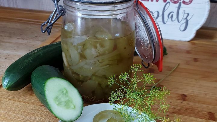 Ein Glas eingelegter Senfgurken steht neben frischen Gurken und einem Teller mit Gurkenscheiben auf einem Holzbrett.