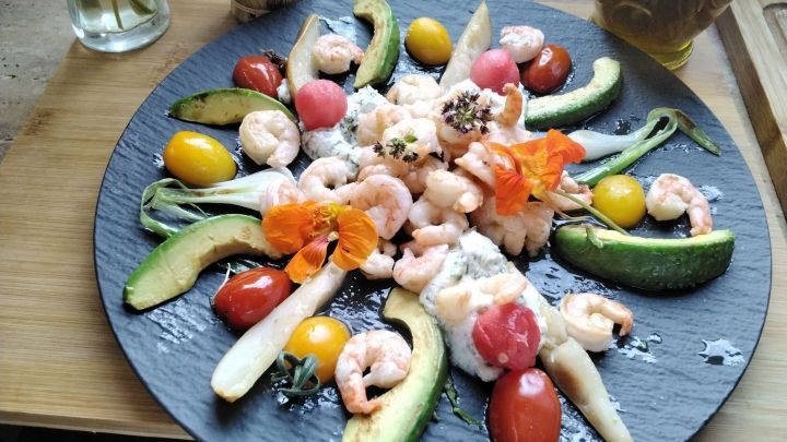 Ein farbenfroher Salat auf einem schwarzen Teller, dekoriert mit Black Tiger Garnelen, Burrata und frischem Gem&uuml;se.