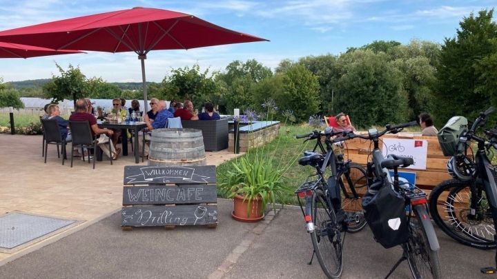 Ein Weincaf&eacute; mit G&auml;sten unter roten Schirmen, Fahrr&auml;der parken daneben, umgeben von Weinbergen und Natur.