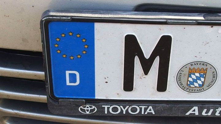 Ein schmutziges Nummernschild mit dem Buchstaben M zeigt, wie Rasierschaum Fliegendreck entfernt. Es ist an einem Toyota befestigt.