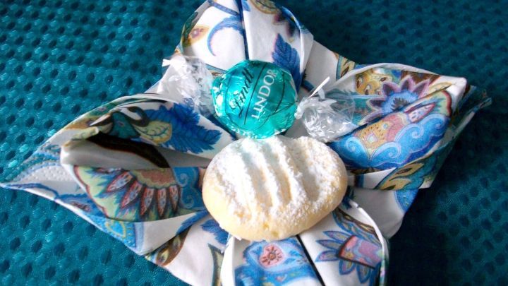 Eine kunstvoll gefaltete Serviettenblume pr&auml;sentiert s&uuml;&szlig;e Leckereien wie einen Keks und eine Praline auf einem blauen Tuch.