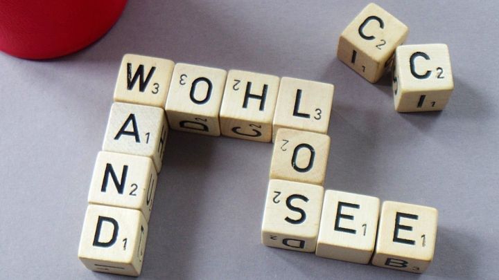 Holzbuchstaben formen W&ouml;rter wie "WAND", "WOHL" und "LOSE" auf einer grauen Oberfl&auml;che, unterst&uuml;tzt von einem roten Becher.