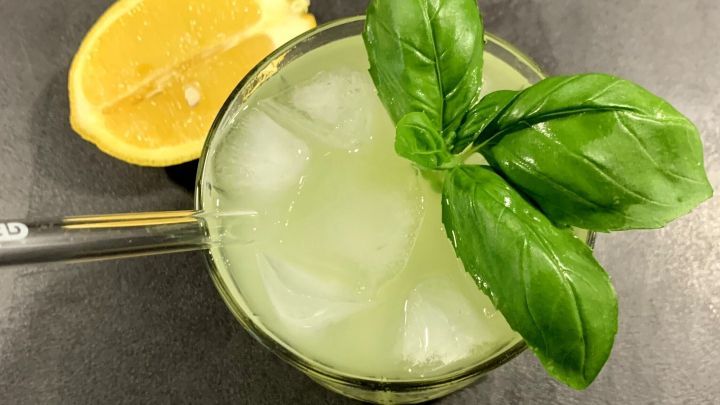 Ein frischer Gin Basil Smash mit Eisw&uuml;rfeln und einem gro&szlig;en Basilikumblatt, neben einer halbierten Zitrone auf dem Tisch.