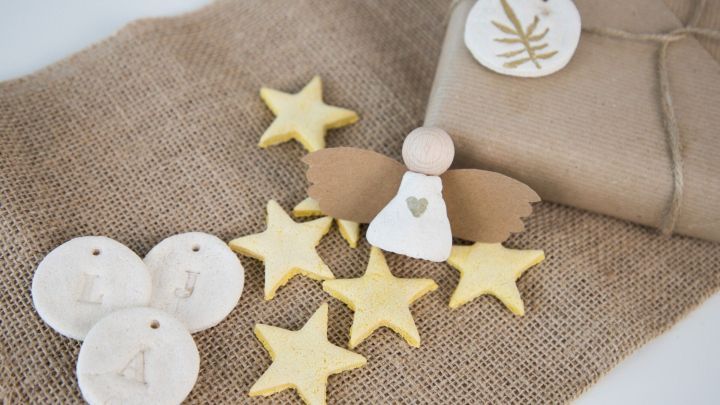 Gelbe Salzteigsterne und ein Engel aus Holz liegen auf einem juten Stoff neben einem liebevoll verpackten Geschenk.