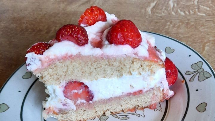 Ein St&uuml;ck Erdbeertorte mit Sahne und frischen Erdbeeren liegt auf einem dekorierten Teller auf einem Holztisch.
