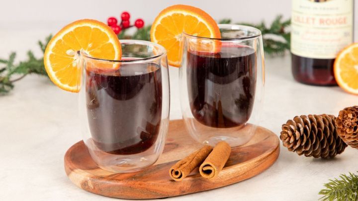 Zwei Gl&auml;ser mit Lillet Hot Berry, gef&uuml;llt mit dunklem Getr&auml;nk, serviert auf einem Holzbrett mit Orangen und Zimtstangen.