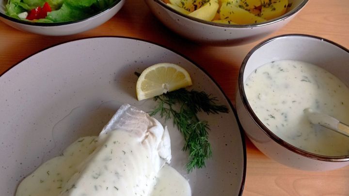 Zarter Kabeljau liegt in Dill-Sahne-B&eacute;chamel, umgeben von Salat und Kartoffeln, alles auf einem Holzuntergrund serviert.