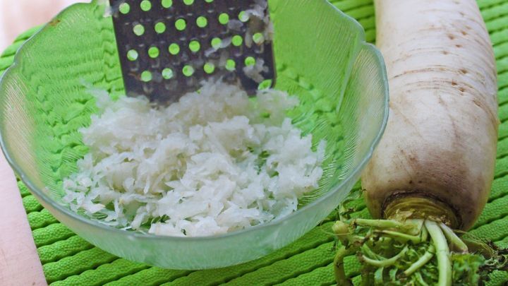 Ein frischer Meerrettich wird gerieben und landet in einer transparenten Sch&uuml;ssel, umgeben von einer gr&uuml;nen Unterlage.