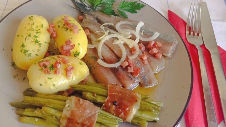 Ein Teller zeigt Matjes in Speckso&szlig;e, umgeben von Kartoffeln und gr&uuml;nem Spargel, angerichtet auf einem karierten Tisch.