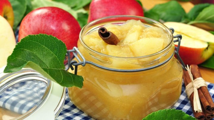 Ein Glas Apfelmus steht im Vordergrund, umgeben von frischen &Auml;pfeln und Zimtstangen, auf einem karierten Tisch.