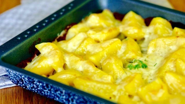 Ein goldbraun &uuml;berbackenes Kartoffelgratin liegt in einer blauen Auflaufform auf einem Holztablett, umgeben von einer Tischdecke.