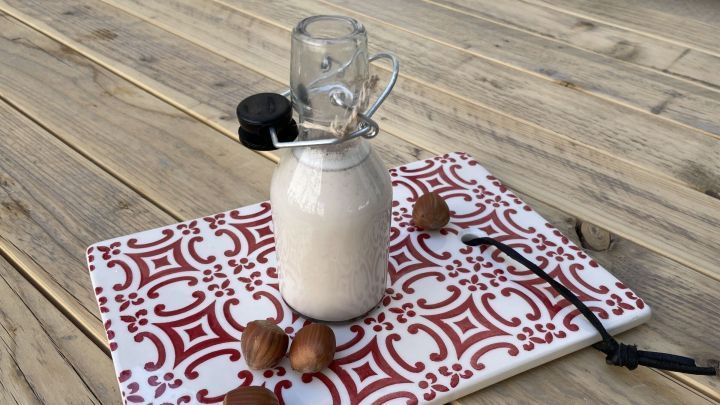 Ein Glas mit Haselnussmilch steht auf einem dekorativen Brett, umgeben von Haseln&uuml;ssen auf einem Holztisch.