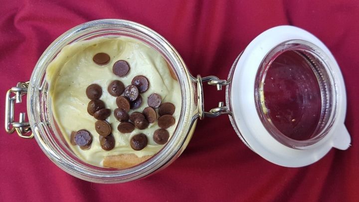Ein Glas Tiramisu mit K&uuml;rbiskern&ouml;l, bedeckt mit Schokoladenst&uuml;ckchen, steht auf rotem Stoff.