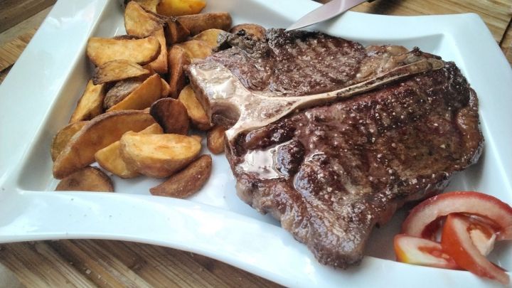 Saftiges T-Bone Steak liegt auf einem wei&szlig;en Teller neben knusprigen Wedges, eingebettet in eine rustikale K&uuml;che.