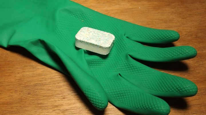 Eine gr&uuml;ne Gummihandschuhfl&auml;che h&auml;lt einen Geschirrsp&uuml;lreinigertab, bereit, den Backofen zu reinigen.