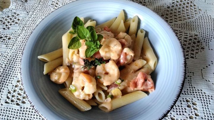 Zarte Garnelen in w&uuml;rziger So&szlig;e liegen auf Penne-Nudeln, dekoriert mit frischen Kr&auml;utern auf einem Tisch mit Spitzenunterlage.