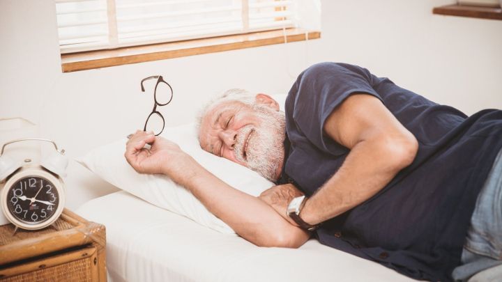 Ein &auml;lterer Mann schl&auml;ft gl&uuml;cklich im Bett, seine Brille liegt neben ihm, w&auml;hrend ein Wecker auf dem Nachttisch steht.