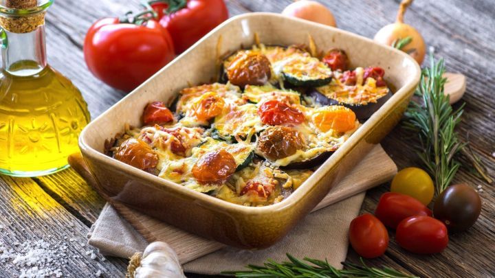 Ein Auberginenauflauf ist mit Tomaten und K&auml;se &uuml;berbacken und liegt in einer braunen Auflaufform auf einem rustikalen Holztisch.