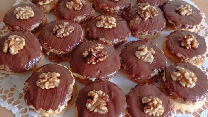 Auf einem filigranen Tuch liegen viele runde Nuss-Doppeldecker, die mit Schokolade &uuml;berzogen und mit Waln&uuml;ssen garniert sind.