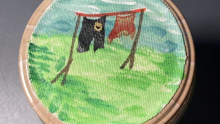 Ein Schraubglas mit einem bemalten Deckel zeigt W&auml;sche auf der Leine, umgeben von einer gr&uuml;nen Wiese und blauem Himmel.