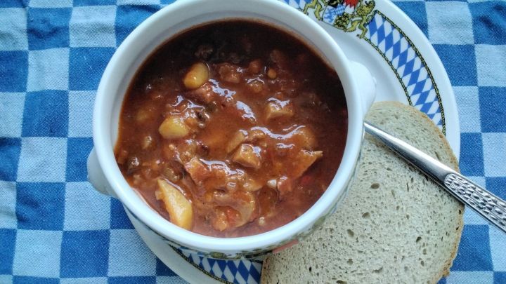 Eine Sch&uuml;ssel Gulaschsuppe mit Kartoffeln und Fleisch steht auf einem Teller, erg&auml;nzt durch ein St&uuml;ck Brot auf einem karierten Tuch.