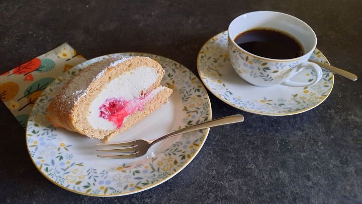 Ein St&uuml;ck Himbeerroulade aus Buchweizenmehl liegt auf einem blumigen Teller neben einer Tasse Kaffee auf einem dunklen Tisch.