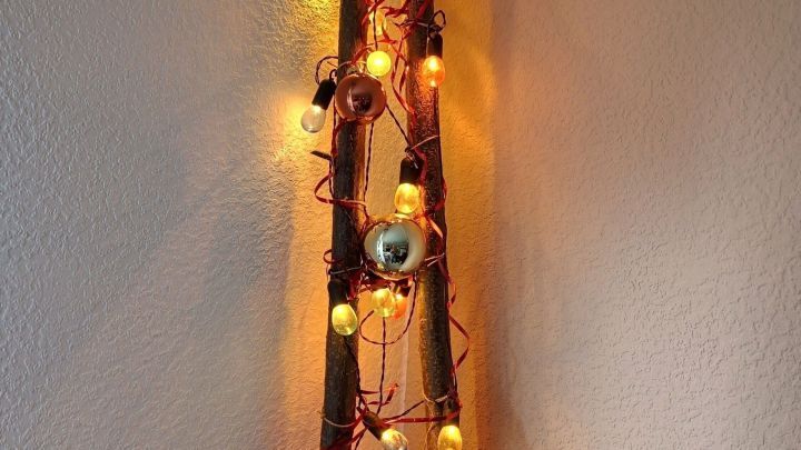 Ein rustikales Dekost&uuml;ck aus zwei &Auml;sten, dekoriert mit bunten Lichtern und Weihnachtskugeln, steht an der Wand.