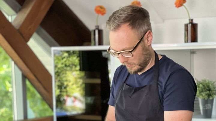 Ein Koch schneidet eine gelbe Paprika auf einem Holztisch, umgeben von frischem Gem&uuml;se in einer modernen K&uuml;che.