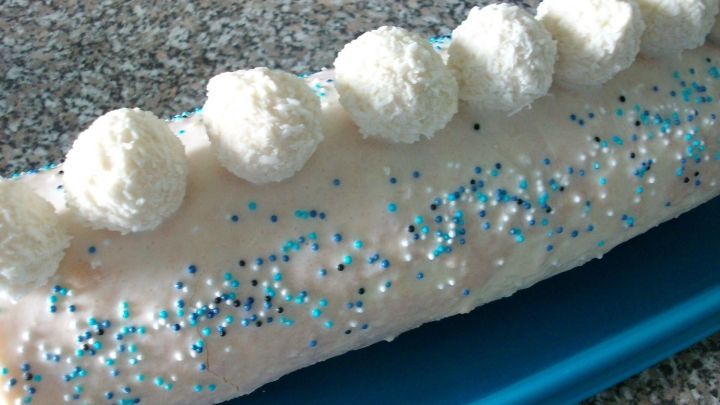 Ein l&auml;nglicher Raffaello-Rolle gleitet auf einem blauen Teller, dekoriert mit Zuckerstreuseln und Kokoskugeln.