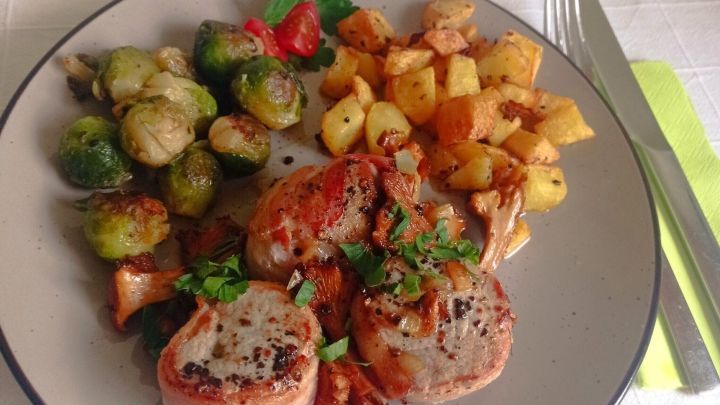 Schweinefilet im Speckmantel liegt appetitlich angerichtet auf einem Teller, begleitet von Bratkartoffeln und Rosenkohl.