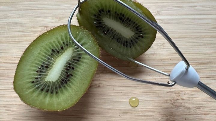 Eine halbierte Kiwi liegt auf einem Holzbrett, w&auml;hrend ein R&uuml;hrger&auml;t daneben liegt und ein Tropfen Saft sichtbar ist.