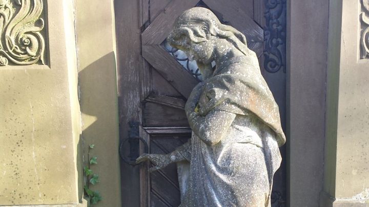 Eine Statue einer trauernden Frau steht vor einer alten Holzt&uuml;r in einem gepflegten Grabbereich, umgeben von Efeu und herbstlichen Bl&auml;ttern.