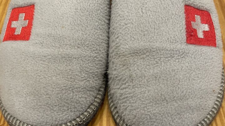 Graue Filzpantoffeln liegen auf einer h&ouml;lzernen Oberfl&auml;che, mit roten Schweizer Kreuz-Emblemen auf jeder Seite.