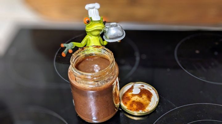 Ein Glaskrug mit Winzer-Marmelade wird von einer niedlichen gr&uuml;nen Fruchtfarbfigur mit einer Kochm&uuml;tze und einem L&ouml;ffel dekoriert, auf einem Tisch.