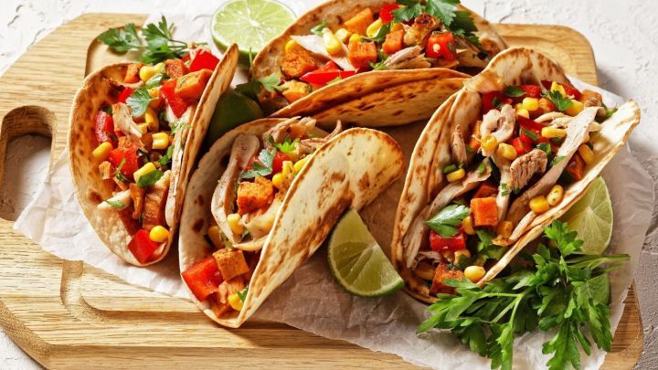 Frische Tacos aus Tortillas mit H&auml;hnchen, Gem&uuml;se und Limette auf einem Holzbrett in heller Umgebung.