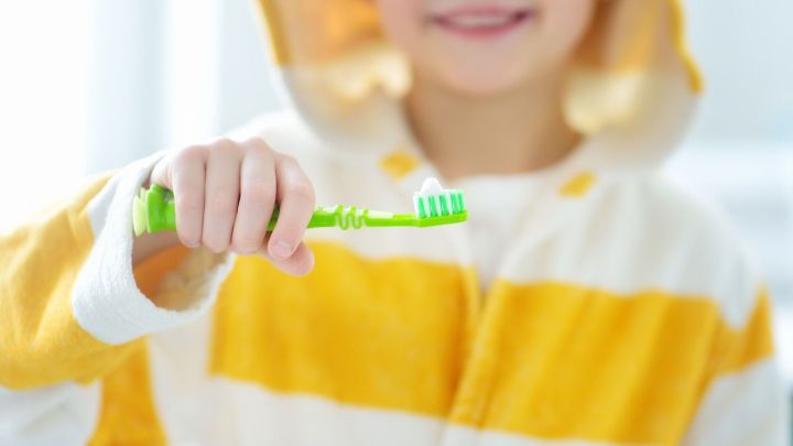 Ein M&auml;dchen in einem gestreiften Bademantel h&auml;lt eine gr&uuml;ne Zahnb&uuml;rste mit Zahnpasta in einem hellen Badezimmer.