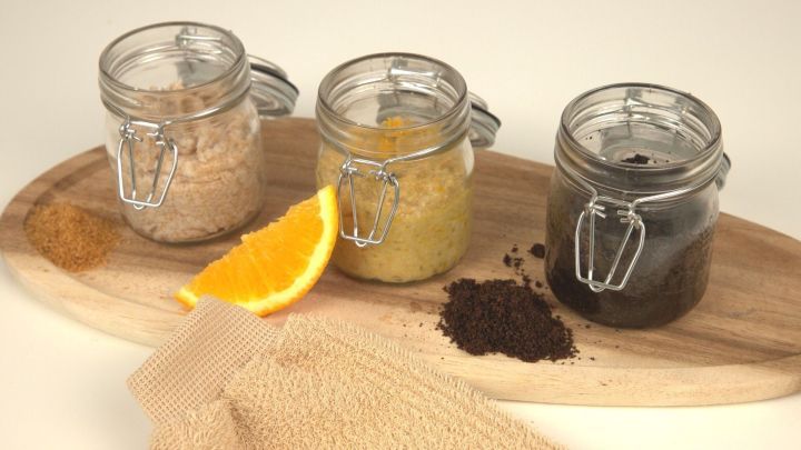 Drei Glasbeh&auml;lter mit K&ouml;rperpeeling, orangefarbener Frucht und einem Peelinghandschuh auf einem Holzbrett.