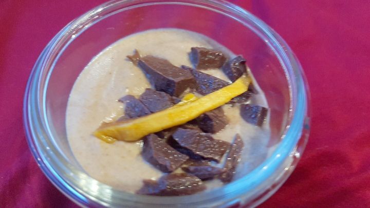 Eine cremige Lebkuchenmousse in einem Glas, garniert mit Schokoladenst&uuml;ckchen und einer Zitronenscheibe auf rotem Tuch.