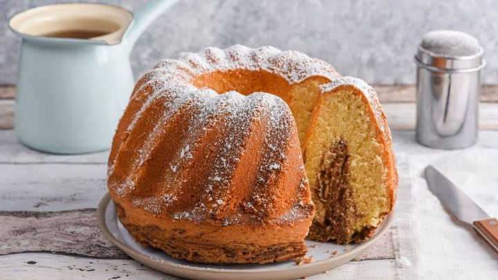 Ein Zimtkuchen in einer Gugelhupfform mit Puderzucker, auf einem Teller, daneben eine Kanne Tee und Zuckerstreuer.