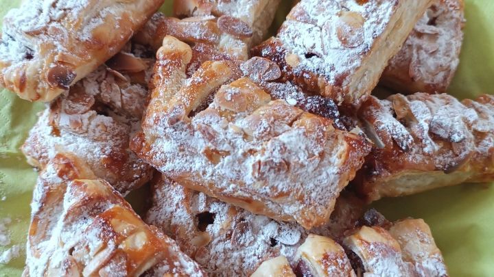 K&ouml;stliche Kirsch- und Apfeltaschen liegen auf einem gr&uuml;nen Serviettenpapier, staubig mit Puderzucker, in einer hellen Umgebung.