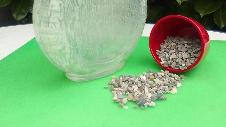 Eine Glasflasche steht auf einem gr&uuml;nen Untergrund, daneben liegt ein roter Becher mit kleinen Steinen.