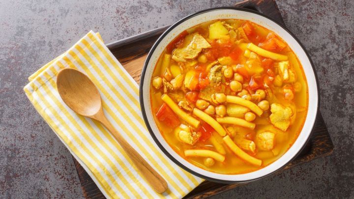 Ein buntes, herzhaftes Rancho Canario f&uuml;llt eine wei&szlig;e Sch&uuml;ssel mit Gem&uuml;se und H&uuml;hnchen, umgeben von einem Holzbrett und einem L&ouml;ffel.