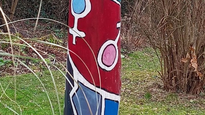 Bunte Holzskulptur steht im Garten und verleiht der Umgebung durch ihre Farben und Symbole eine k&uuml;nstlerische Note.